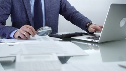 Businessman wearing blue suit is analyzing financial reports with magnifying glass and using calculator in a professional office setting. Low lighting, close-up view. Business people and audit concept