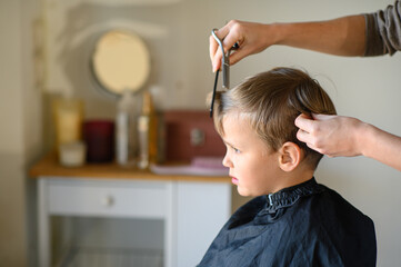 Fototapeta premium A little blond boy gets a haircut, sitting in a salon chair with a cape. The hairdresser holds scissors and a comb. The child is calm and enjoying the experience. Concept of childhood grooming and sel