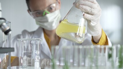 Woman scientist researcher wearing white protective gloves is shaking up a yellow chemical solution inside an Erlenmeyer flask in a laboratory setting, portrait view. Science and medicine