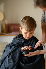 A little blond boy sits in a salon chair while the hairdresser shows him a hair clipper, letting...
