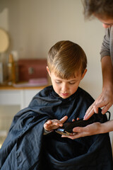 A little blond boy sits in a salon chair while the hairdresser shows him a hair clipper, letting...