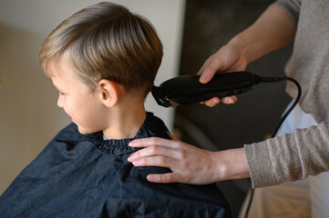 A little blond boy gets a haircut with a hair clipper, sitting in a salon chair with a cape. The...