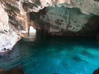 Inside a rugged coastal cave, turquoise water shimmers under the rocky ceiling, revealing the formation’s rough, damp textures and the hidden entrance to the open sea beyond. Light seeps inside.