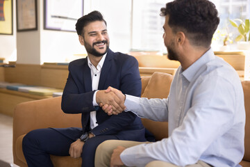 Two millennial 30s businessmen shaking hands, with joyful expression, feel satisfied with teamwork...