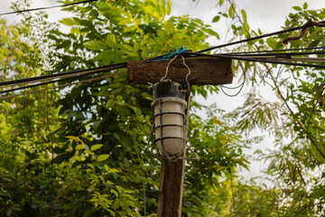 A vintage industrial outdoor lamp with protective metal cage, mounted on a weathered wooden post with electrical wires. Lush green foliage in the background.