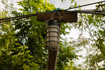 A vintage industrial outdoor lamp with protective metal cage, mounted on a weathered wooden post with electrical wires. Lush green foliage in the background.