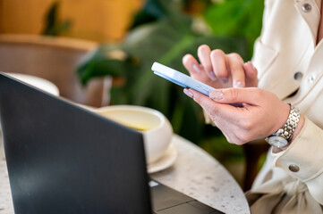 A person using a smartphone while sitting at a table with a laptop and a cup. The background features lush green plants, creating a relaxed workspace atmosphere.