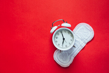 White alarm clock and menstrual sanitary pad on red background, flat lay.