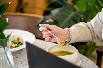 A close-up of a hand holding a spoon with colorful vegetable soup, set against a background of greenery and a laptop, capturing a moment of dining and productivity.