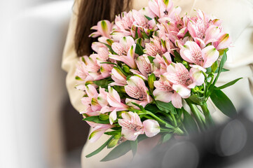 A close-up of a person holding a vibrant bouquet of pink alstroemeria flowers, showcasing delicate petals and rich green leaves, set against a soft, blurred background.