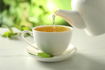 Pouring freshly brewed tea from teapot into cup at white marble table, closeup