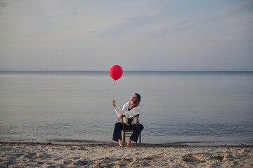 Young woman sits on chair at serene beach, holding vibrant red balloon. Sunset casts peaceful glow...