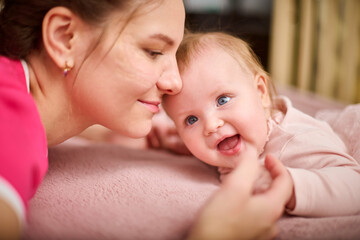 Young woman with brown hair and smiling baby girl on pink bed. Light-hearted interaction, creating joyful atmosphere. Warm lighting enhances bond
