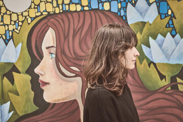 Young woman, brown hair, adult, stands beside vivid mural outdoors. Mural displays artistic portrait and floral patterns, rich in color and stylized design