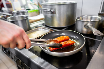 A chef uses tongs to flip salmon fillets sizzling in a frying pan on a commercial kitchen stove. Pots and pans surround the chef, indicating a busy cooking environment.