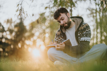 A thoughtful man writing in a notebook while sitting on the grass in sunlight. The serene outdoor...