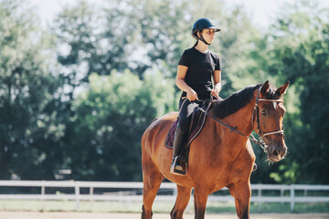 Fototapeta premium A young woman enjoys horseback riding on a sunny day, showcasing her equestrian skills in a peaceful outdoor arena with lush greenery surrounding her.