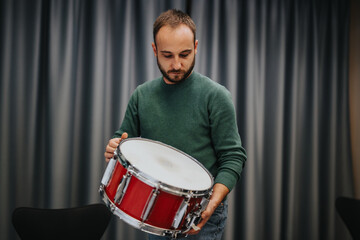A man in a green sweater closely examines a red drum in a studio with curtains.