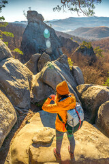 A female hiker in a yellow jacket and beanie checks her wristwatch on a rocky trail, surrounded by...