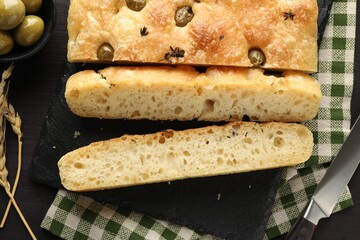 Pieces of delicious focaccia bread with olives, thyme, spikes and knife on dark wooden table, flat lay
