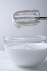 Hand mixer with whisks and bowl of whipped cream on white wooden table, closeup