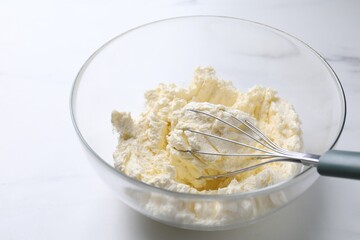 Bowl with whisk and whipped cream on white marble table, closeup