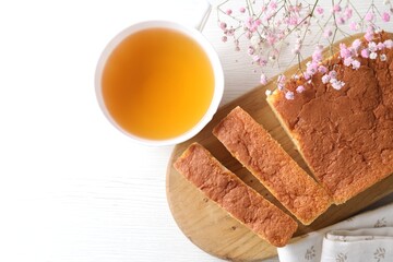 Cut tasty Japanese Castella sponge cake, tea and flowers on white wooden table, flat lay. Space for...