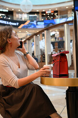 A young woman of European appearance is talking on the phone over a glass of coffee at a table in a shopping center cafe