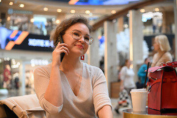A young woman of European appearance is talking on the phone over a glass of coffee at a table in a shopping center cafe