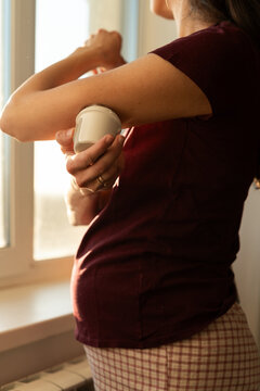 Close-up of a pregnant woman standing by a window attaching a continuous glucose monitoring on her arm