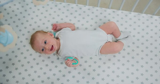 Close up infant playing with teether of teeth in crib, top view Portrait of baby boy lying on back and actively moves his arms and legs in baby cot.