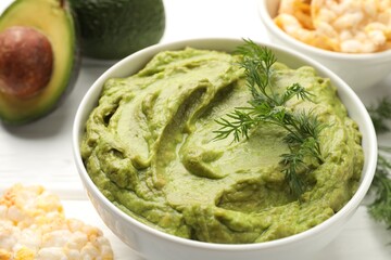 Tasty avocado dip in bowl, dill and puffed rice cakes on white table, closeup