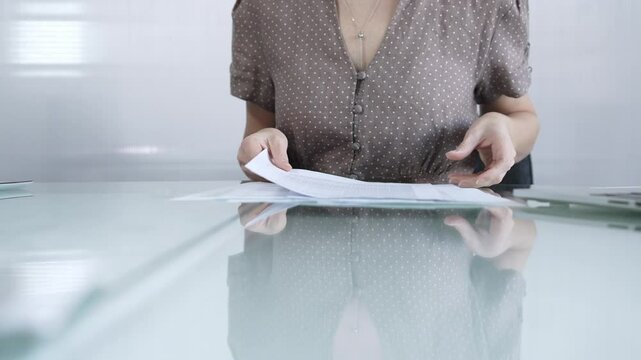 Businesswoman or accountant in brawn dotted blouse is going through paperwork while holding documents at a glass desk in a well lit, contemporary office setting. Business people concept