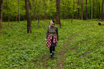 Young female hiker with backpack and plaid shirt tied around her waist enjoys a peaceful walk through a vibrant green forest, embracing the tranquility of nature during her outdoor adventure