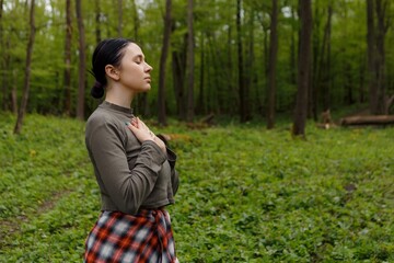 Young woman standing in verdant forest, palms resting on chest, experiencing peaceful moment while...