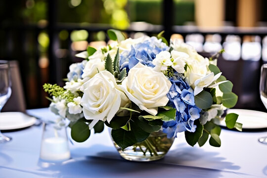 Beautifully wedding bouquet featuring white roses and blue hydrangeas with lush greenery in clear glass vase. Elegant floral arrangement on table, enhancing romantic ambiance of wedding celebration