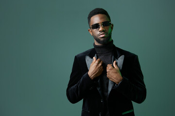 Confident man in sunglasses and black tuxedo jacket standing against vibrant green background, exuding sophistication and style