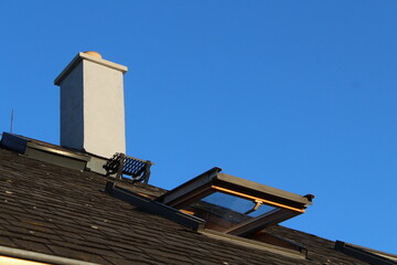 Open skylight window and chimney on a black roof against blue sky background 
