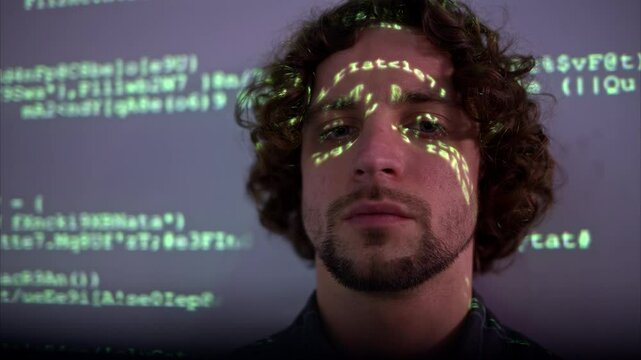 Man with curly hair in front of a projection of green computer code, illuminating on his face