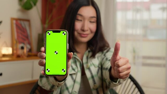 Asian Korean Chinese Japanese woman showing at camera screen mobile phone smartphone chroma key green display female girl thumb up recommend advertising app mock up businesswoman business home indoors