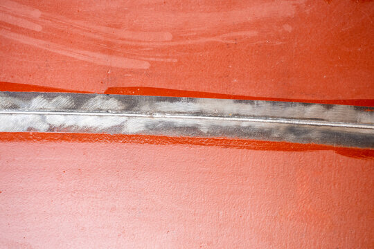In the metalworking industry, a welded seam on a red-painted metal surface is shown, with visible grinding marks and a smooth, even weld bead