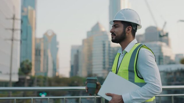 Side view portrait of engineer with laptop and coffee cup walking along bridge against skyscrapers. Bearded contractor in safety vest carries computer to workplace outdoors