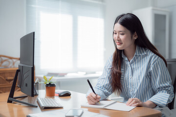 Young Asian businesswoman writing notes in a notebook while focused on a computer screen, working...