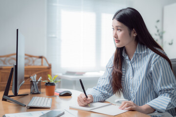 Young Asian businesswoman taking notes in her notebook while focused on the computer screen, working efficiently from her cozy home office during the day, enjoying natural light