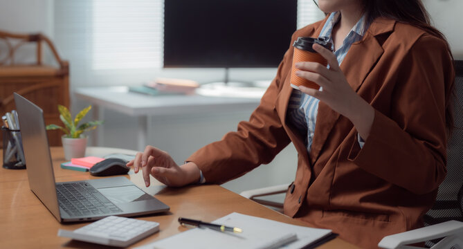 Businesswoman wearing brown jacket and striped shirt drinking coffee and working on laptop in bright modern office, using touchpad and keyboard