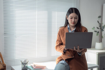 Young Asian businesswoman sitting near window and using laptop while working from home office with...