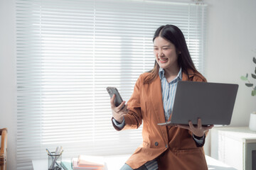 Young Asian businesswoman comparing information on smartphone and laptop while smiling in modern...