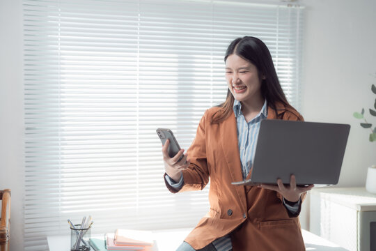 Young Asian businesswoman comparing information on smartphone and laptop while smiling in modern office, efficient multitasking professional managing business operations