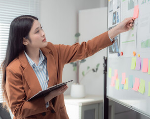 Young businesswoman holding a clipboard and attaching colorful sticky notes on a whiteboard while presenting ideas in a modern office environment, engaging her team in strategic planning