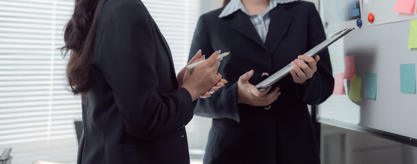 Two businesswomen standing next to a whiteboard with sticky notes, discussing work and holding a clipboard and a pen, in a bright office environment